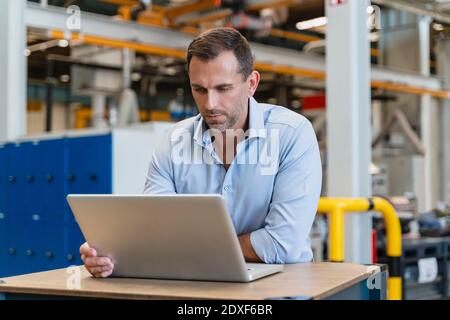 Male entrepreneur using laptop while leaning on desk in factory Stock Photo
