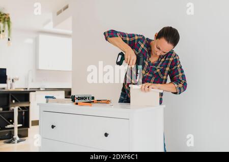 Craftsperson using electric screwdriver while standing by cabinet at home Stock Photo