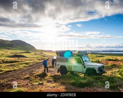 Sun setting over lone traveler standing behind off-road vehicle and waving at camera, Iceland Stock Photo