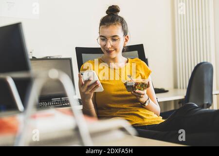 Smiling businesswoman using mobile phone during lunch break at office Stock Photo