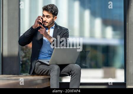 Angry businessman talking on mobile phone while sitting with laptop outdoors Stock Photo
