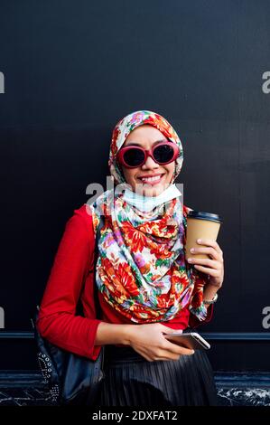 Smiling Muslim woman holding mobile phone and coffee cup against black wall during COVID-19 Stock Photo