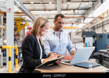 Female entrepreneur with book while male colleague working on laptop in factory Stock Photo