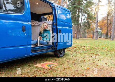 Thoughtful woman holding tea cup while sitting at door of camper van in Cannock Chase Stock Photo