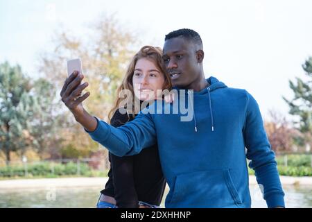 African black man and redhead caucasian woman smiling and taking a selfie with their smartphone in a park. Young multiracial couple portrait. Stock Photo