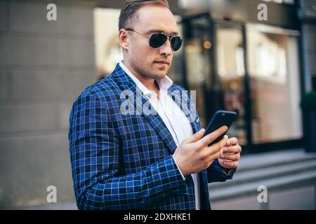 Closeup Portrait of stylish handsome young man standing outdoors. Man wearing jacket and shirt. man using mobile phone Stock Photo