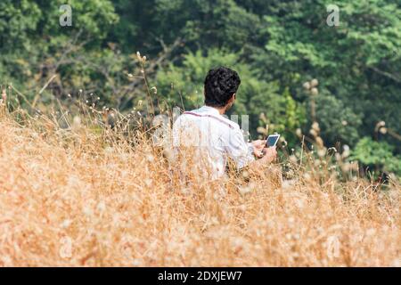 A young man with a smart cell phone  sitting in the dry grass flowers  in autumn on the top of mountains of Sanjay Gandhi National Park, Mumbai, India Stock Photo