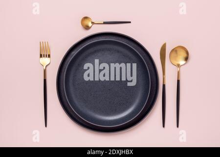 Dark ceramic plates and gold and black cutlery on pastel pink background. Top view - Image Stock Photo