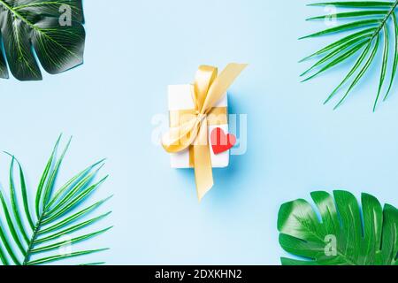Valentines' day concept. White gift box postcard with a gold bow ribbon and Tropical jungle Monstera green leaves and red heart isolated on blue backg Stock Photo