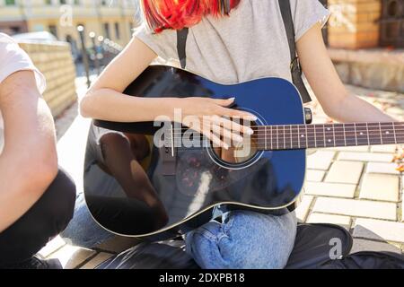 Girl with colored hair singing and playing acoustic guitar. Teenagers sitting on sidewalk on city street