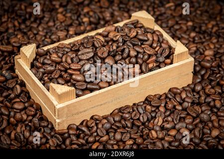 Closeup of roasted coffee beans in a small wooden crate over a pile of coffee beans. Stock Photo