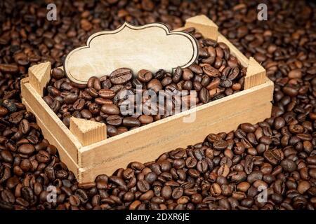 Closeup of roasted coffee beans in a small wooden crate with empty label, over a heap of coffee beans. Stock Photo