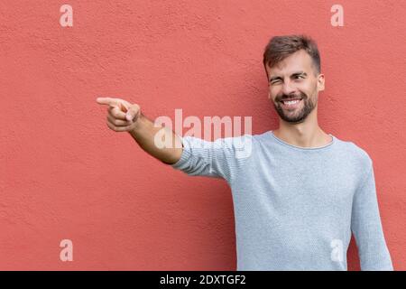 Handsome young Caucasian bearded man in grey shirt smiling and pointing finger to camera right on orange painted wall background with free space for t Stock Photo