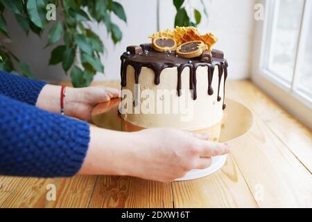 Woman's hands putting a caramel cake decorated with chocolate and waffles on wooden table near the window. Stock Photo