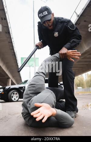 low angle view of aggressive police officer with truncheon arresting hooded offender lying on urban street Stock Photo