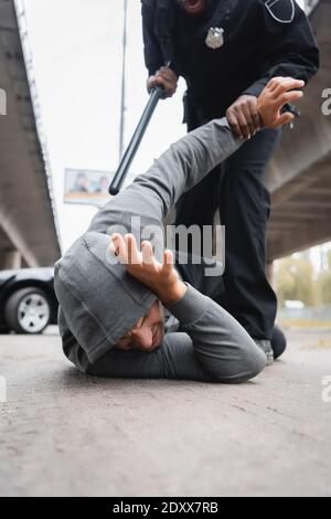 african american police officer with truncheon arresting hooded thief lying on street on blurred background Stock Photo
