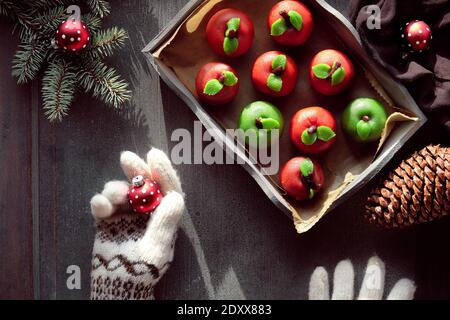 Marzipan sponge apples. Tasty creative dessert on rustic wooden tray with textile cotton towel. Hands in wool gloves. Stock Photo