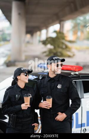 Smiling officers of police in sunglasses holding coffee to go near auto on blurred background on urban street
