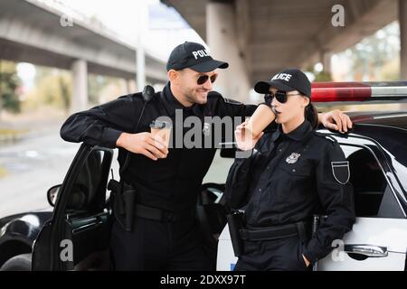 Smiling officer of police in sunglasses looking at colleague drinking coffee to go near car outdoors