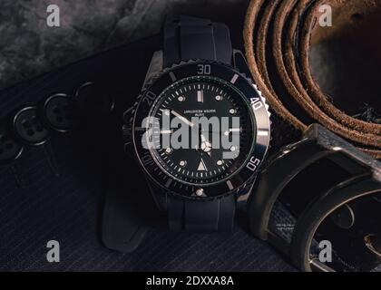 Close-up of a black-chrome watch, a belt buckle of a leather belt on top of part of a sleeve of a jacket Stock Photo