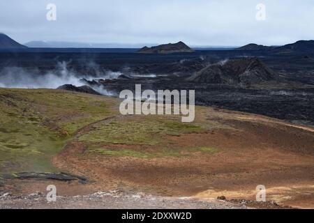 Steam rises from the ground at the Krafla Lava Fields in the Lake Myvatn Area in North Iceland. A volcano cone can be seen rising from the black rock. Stock Photo