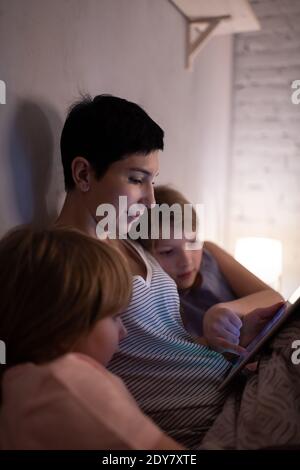Little girls and adult woman playing game on tablet while lying in bed and spending time at home together Stock Photo