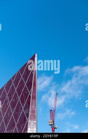 Looking up at shiny red modern and contemporary office building against a blue sky with small clouds and a crane Stock Photo