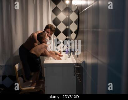Side view of adult male in little boy with shaving foam on cheeks washing hands under running water while spending time together in bathroom Stock Photo