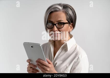 Portrait of a grey haired mature woman in white shirt and eye glasses using digital tablet. The use of technology by the elderly browsing internet Stock Photo