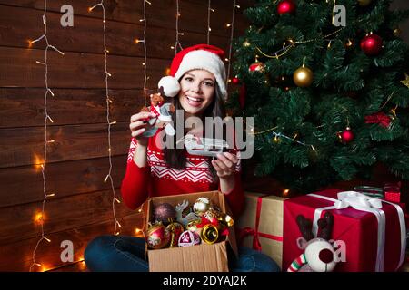 Smiling young woman in Santa hat putting decorations on xmas tree at home Stock Photo