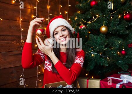 Smiling young woman in Santa hat putting decorations on Christmas tree at home Stock Photo