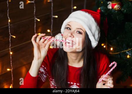 Funny girl in Santa hat with red lips and lollipop candy in her hand on Christmas background. Stock Photo