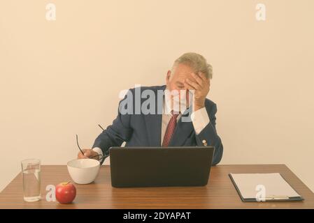 Studio shot of handsome senior bearded businessman holding eyeglasses while having headache with laptop and basic things for work on wooden table Stock Photo