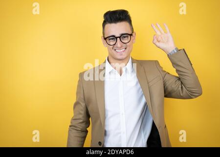 Young business man over isolated yellow background doing ok sign with fingers and smiling, excellent symbol Stock Photo