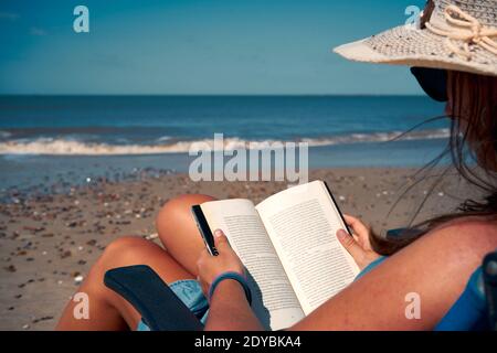 A sun-tanned female in a Panama hat sitting in sunbed and reading a book on the beach, beautiful seascap in the background Stock Photo