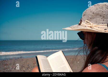 A dark-haired female in a Panama hat reading on the beach, beautiful sea view in the background Stock Photo