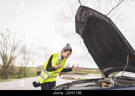 Young Caucasian woman looking at car engine, she doesn't know what to do, car broken down in the middle of the road. Automobile and Roadside Assistanc Stock Photo