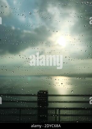 A vertical shot of a window covered in raindrops with the sea under the sunlight on a blurry background Stock Photo
