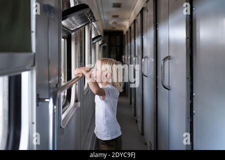 Blond boy looks curiously at the train window. Traveling with children. Stock Photo