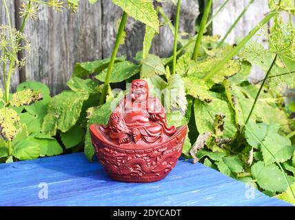 Ceramic buddha figurine on blue aged wooden boards in summer park with flowers. Stock Photo