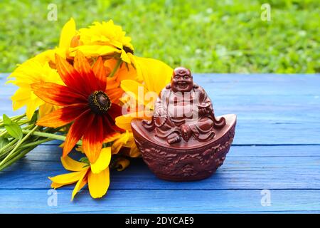 Ceramic buddha figurine on blue aged wooden boards in summer park with rudbeckia flowers. Stock Photo