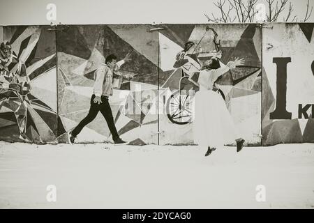 cool young newlyweds in wedding clothes and denim jackets jumping against the background of the wall painted pop art street art in winter in the snow. Stock Photo
