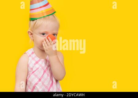 Cute blonde girl in party hat touches your clown nose. Portrait close up on yellow background. Copy space. Stock Photo