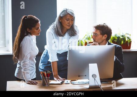 Colleagues discussing a new presentation sitting at coworking room looking at desktop computer. Team on meeting in modern office interior Stock Photo