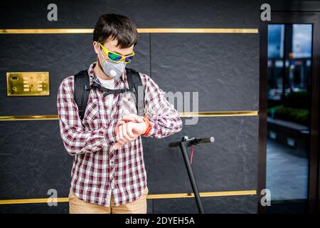 Courier uses electric scooter in city. Young attractive Caucasian man with backpack, stylishly dressed, using mask against smog and air pollution in Stock Photo