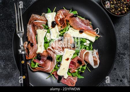 Prosciutto crudo ham salad with brie camembert cheese and arugula on a plate. Black background. Top view Stock Photo