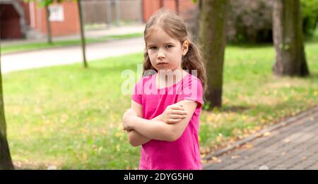 One lone offended child, sad anxious angry lone little girl standing slightly upset, kid with arms crossed, closeup, portrait, outdoors, copy space Stock Photo