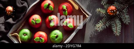 Marzipan sponge apples, panoramic image. Tasty Christmas dessert on rustic wooden tray with textile cotton towel. Hands in wool gloves hold Xmas Stock Photo