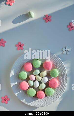 Springtime sweets on white background with long shadows. Marzipan Easter eggs, pink and mint green macaroons on white plate. Tasty creative dessert in Stock Photo