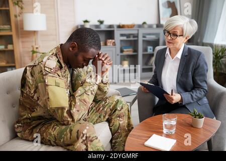 Depressed young military man sitting on sofa during therapy session with psychologist Stock Photo